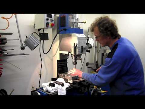 A man in a blue shirt using a drill press in a workshop surrounded by tools hanging on the wall.