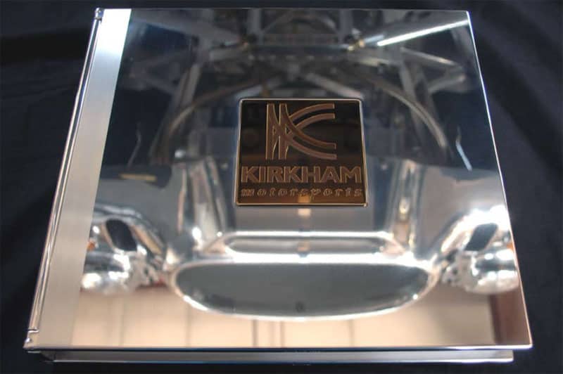 Polished kirkham motorsports metal plate with a reflective surface and engraved logo, displayed over a blurred background of a chrome car structure.