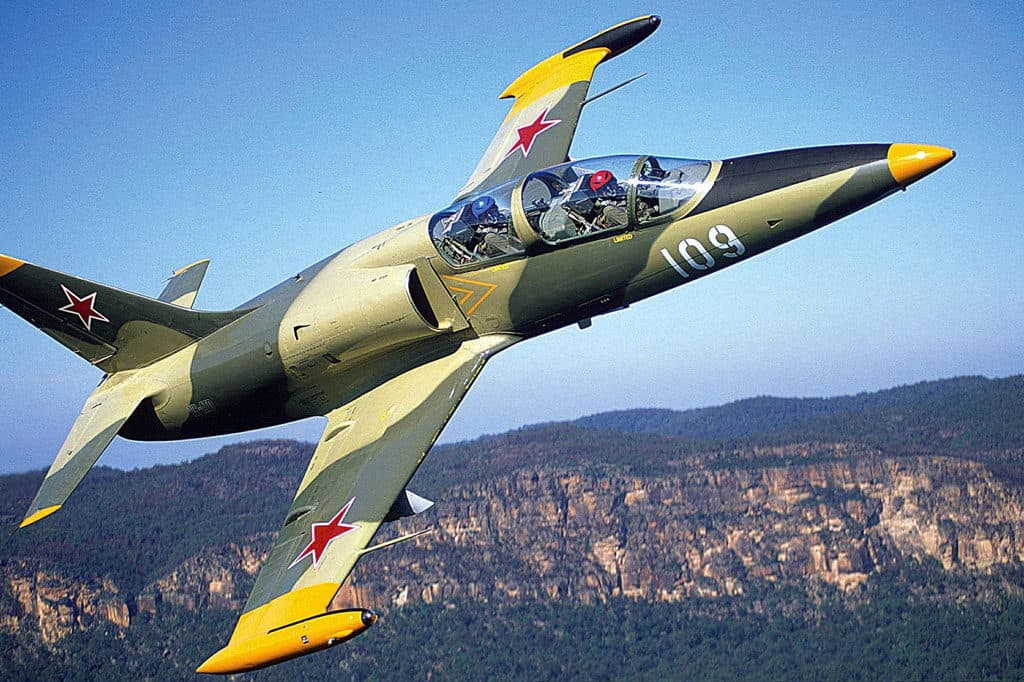 A vintage L-39 Albatros jet fighter in flight against a blue sky, with dark green body, yellow accents, and red stars on its wings.