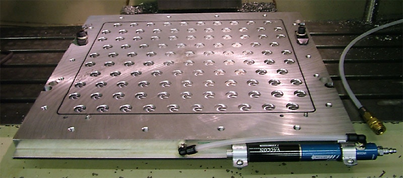 Metallic vacuum table with a grid of holes and a hydraulic piston on the side, situated in an industrial setting.