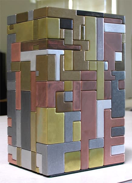 A square-based sculpture featuring an intricate 3d puzzle design with interlocking panels in shades of gold, silver, and bronze.