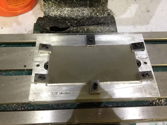A metallic machine component with a rectangular shape, four square corners, two holes, and visible wear.