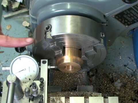 A machinist's hand adjusting a metal part clamped in a lathe, with a dial indicator gauge measuring the part's accuracy.