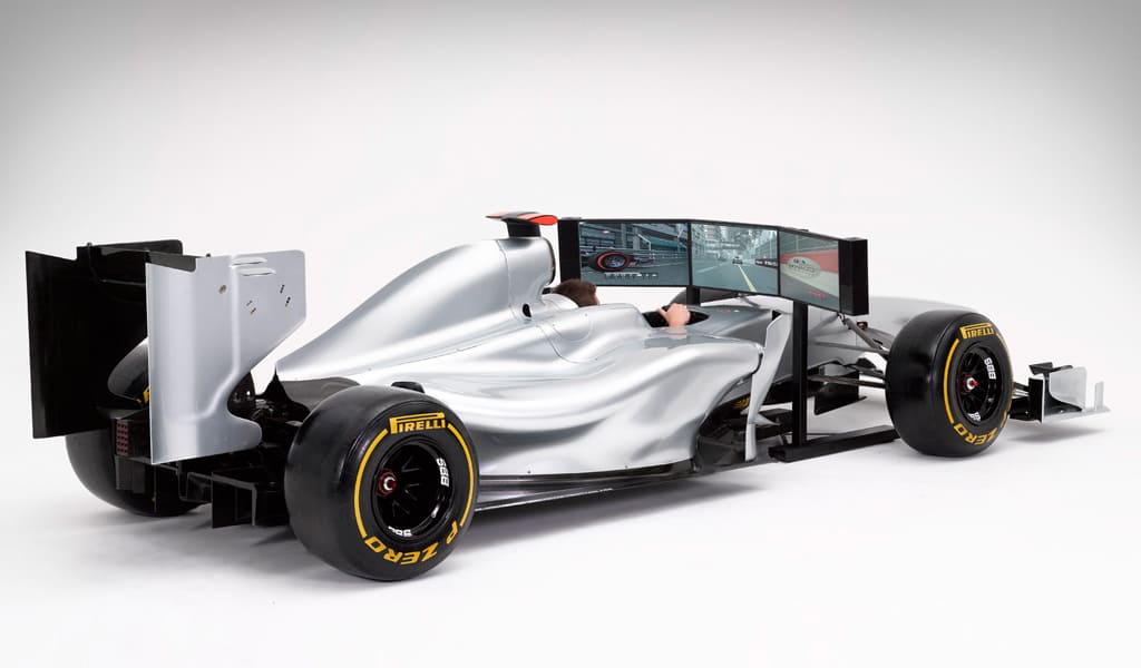 A silver F1 car simulator with sleek design, front wing, rear diffuser, and four side screens.