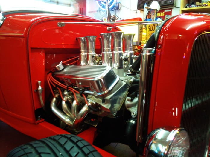 Red vintage car with big block Chevy engine and chrome exhaust pipes, parked near car show signs.