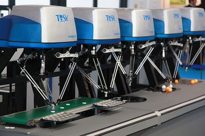 A row of industrial robotic arms with blue and white casings, placed along a conveyor belt handling small objects.