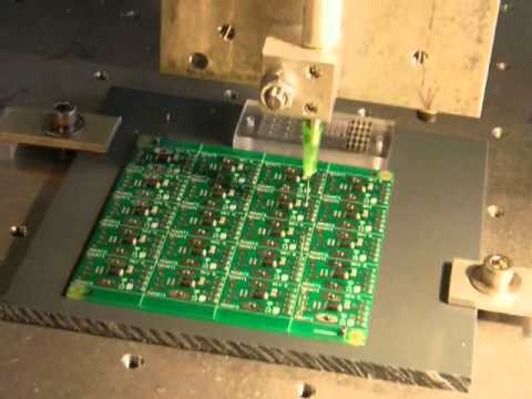 A machine applying solder paste to a green circuit board secured on a metal platform.