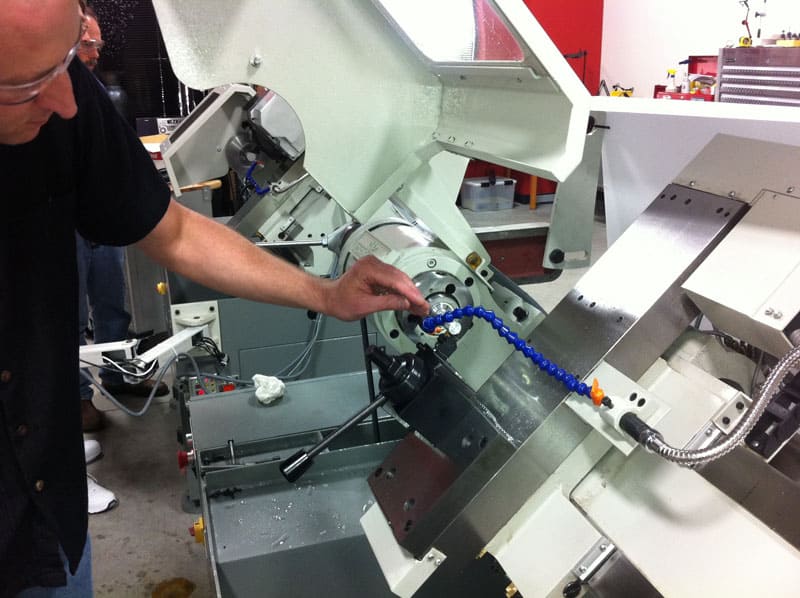 A person wearing safety glasses and a black shirt stands next to an industrial machine with a white body and metal components, featuring a blue chain.