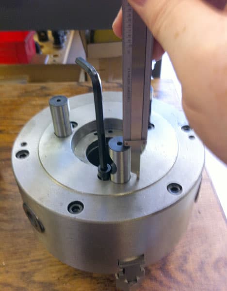 A metal part with holes sits on a wooden table, held by a hand using a caliper against the center hole.