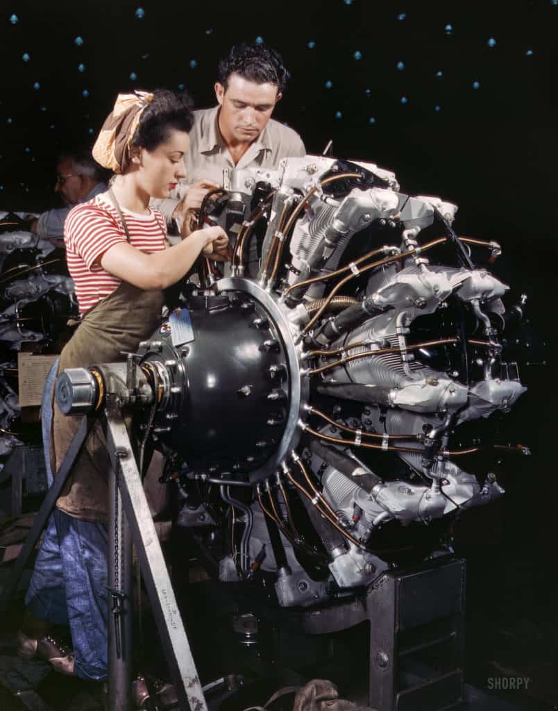 Woman in red-striped shirt working alongside man in tan shirt at an engine in a factory or workshop setting.