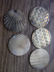 Three silver coin-like objects with intricate guilloche designs and triangular arrangement.