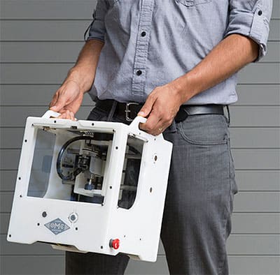 Person in front of slat wall holding white device with plastic window, red button, and "OMC" logo.