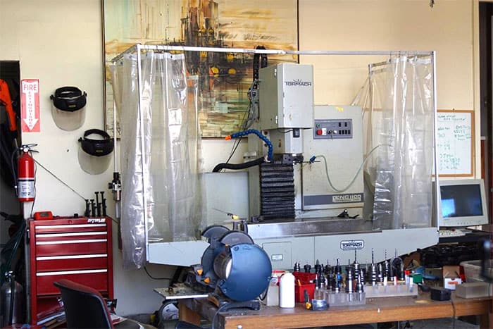 A CNC milling machine in a well-equipped workshop, surrounded by tools, vices, clamps, tool chest, fire extinguisher, and computer equipment.