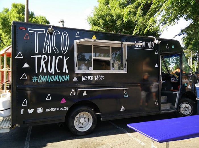 A black food truck with white lettering and geometric shapes parked on a sunny day near a blue tablecloth-covered table.
