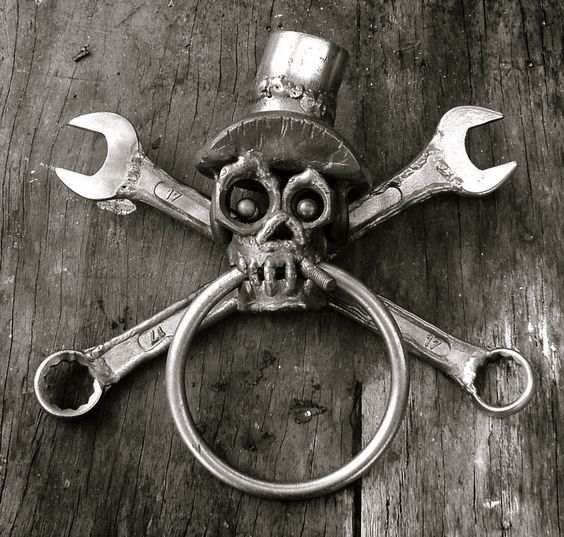 A black and white image of a skull with a top hat, crossed wrenches behind it, and a ring through its nose, arranged on a wooden surface.