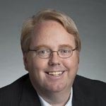 Middle-aged man with short blond hair and black suit jacket, smiling at the camera.