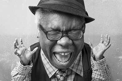 Elderly man in a hat and glasses, yawning or shouting, dressed in a vest and tie, against a textured background.