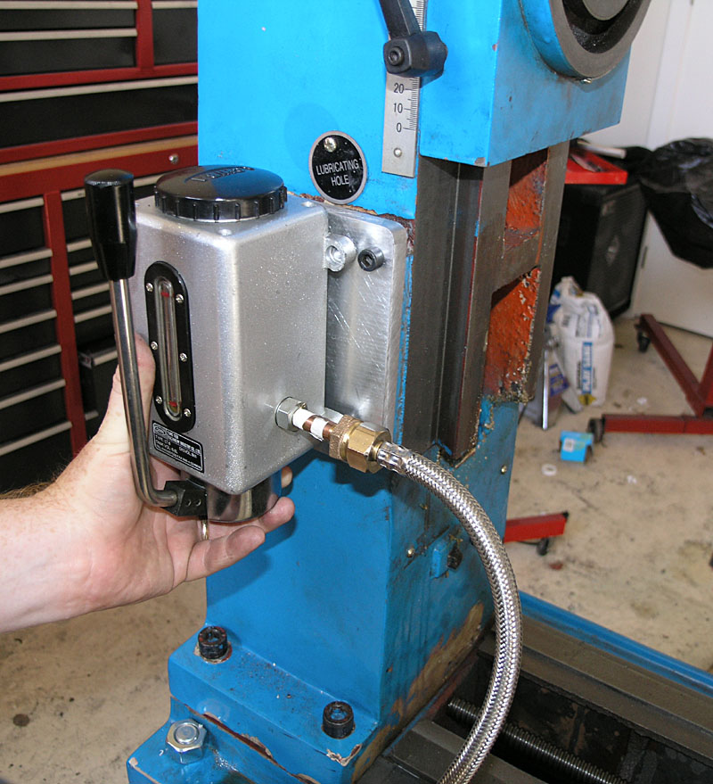 Hand-operated lubrication pump with silver metal body, black handle and knob, supplying oil through flexible hose to machine.
