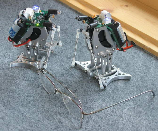 Two robotic penguin-like devices with red wires and circuit boards on top, standing facing each other on a grey surface.