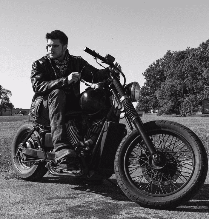 Young man sitting on a motorcycle in a park, wearing jeans and a bandana.