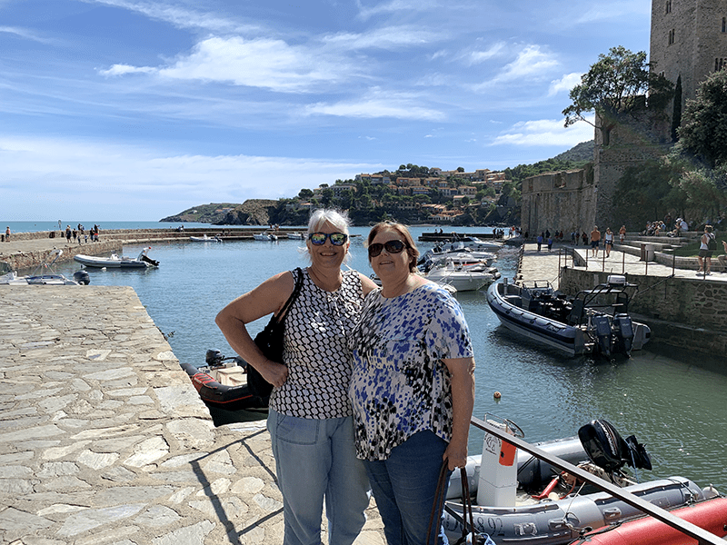 Two women stand on a stone dock overlooking a harbor with boats and surrounding buildings.