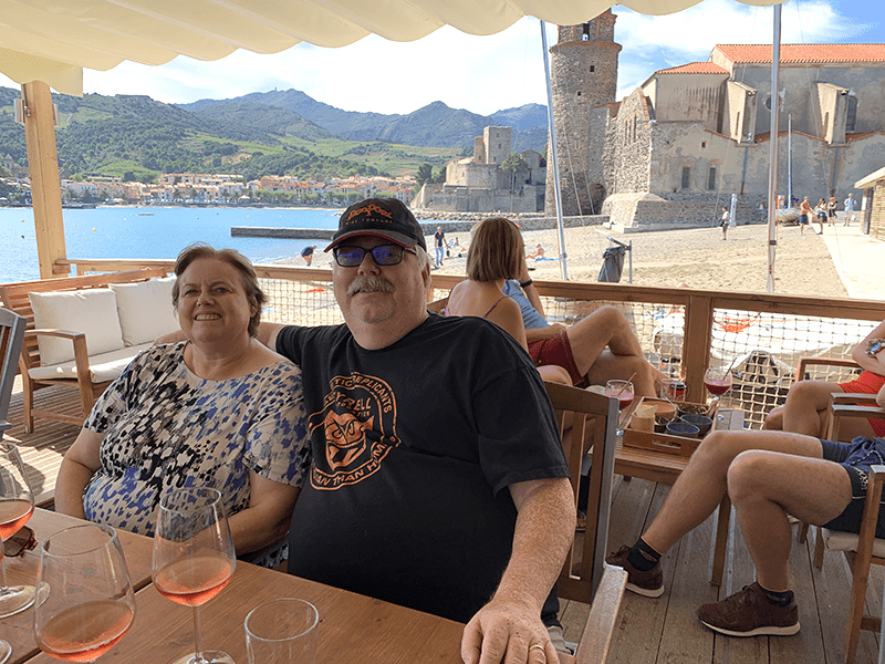 Two people sitting at an outdoor restaurant table overlooking water with mountain scenery in the background.