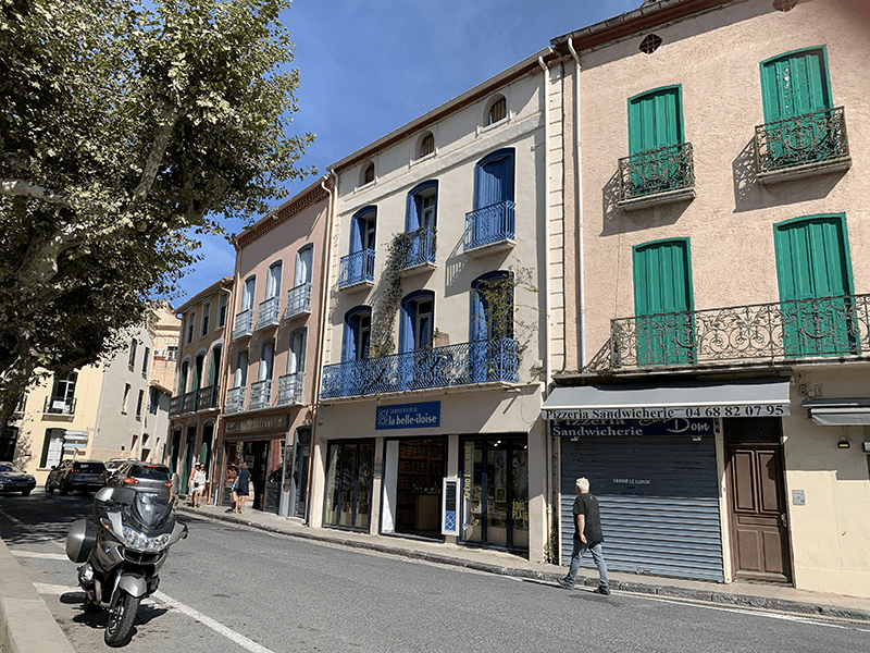 Small town street scene with diverse buildings, beige stucco, blue shutters, green curtains, and a motorcycle.