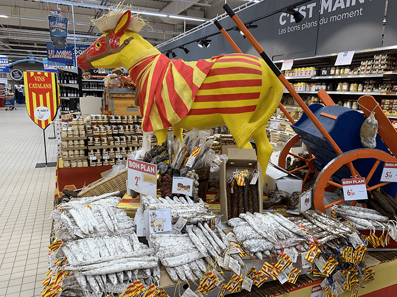 Yellow and red cow statue in grocery store display case surrounded by food products and a "VINS CATALANS" shield.