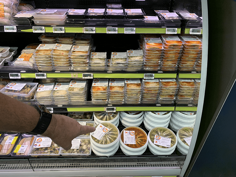 Person browsing refrigerated glass case in grocery store or deli with assorted prepared foods and sandwiches.