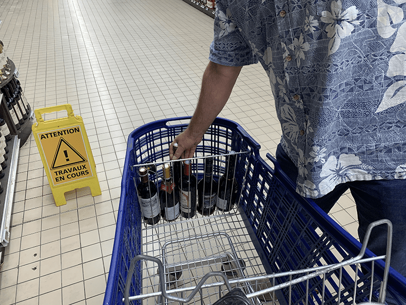 A person in a blue Hawaiian shirt stands next to a blue shopping cart with wine bottles, on a store's tiled floor.