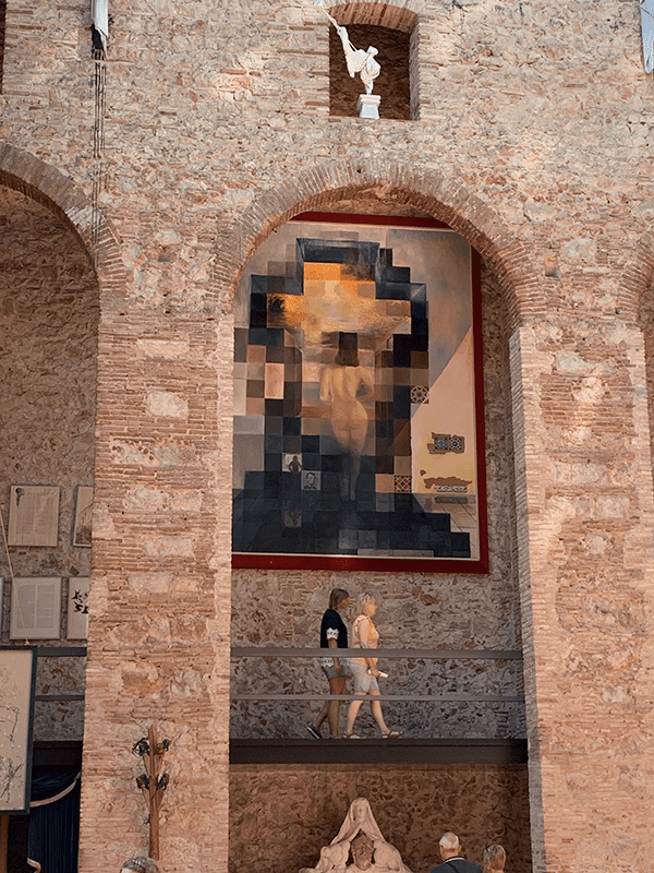 Large stone building with arched doorway and abstract pixelated painting on wall with red border. Two people walk towards the camera.