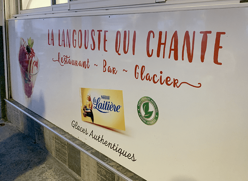 Red sign with yellow background, featuring French text "LA LANGOUSTE QUI CHANTE" and Nestle's La Laitiere logo.