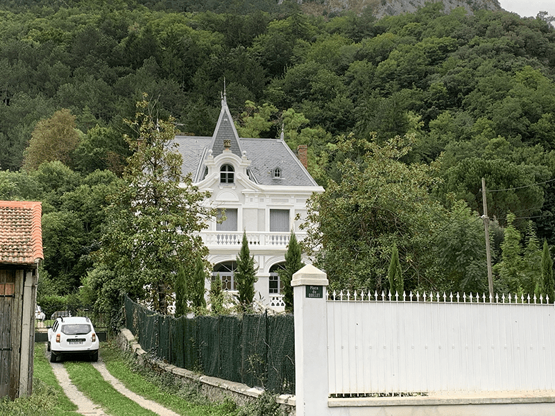 A white mansion with a gray roof, surrounded by lush green trees and tall evergreen trees.
