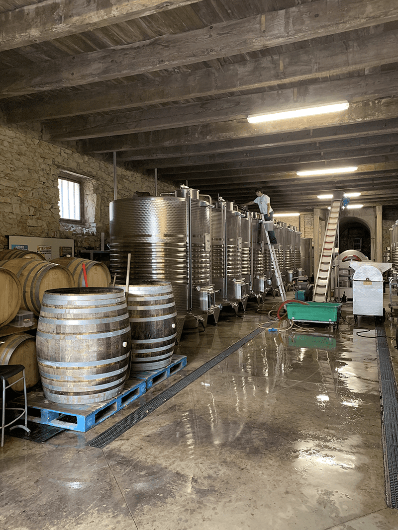 Industrial storage room with metal tanks and wooden barrels, person on ladder in background.