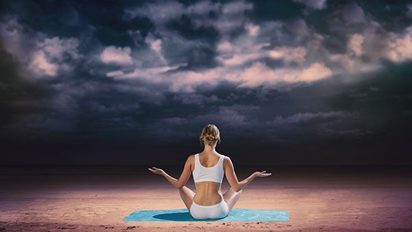 A woman is meditating on the beach, embracing the opportunity for tranquility amid a cloudy sky.