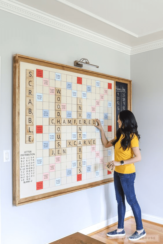 Woman standing in front of a wooden-framed Scrabble board with colored tiles.