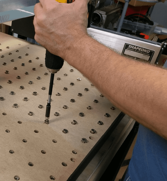 A person's arm holding a drill bit above a workbench with multiple circular holes drilled into it.