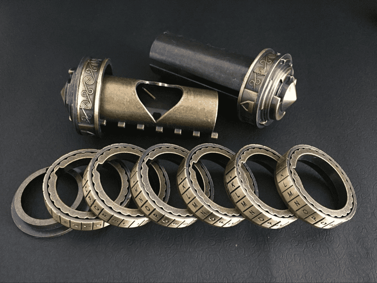 Intricately designed metal rings with engraved letters on a dark gray background featuring two cylindrical objects.