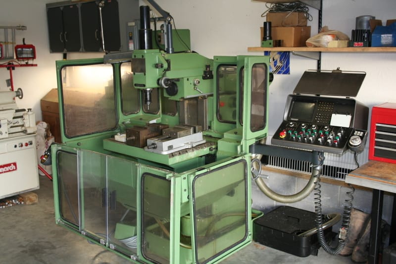 A large green CNC mill in a garage-style workshop with safety glass panels and control panel.