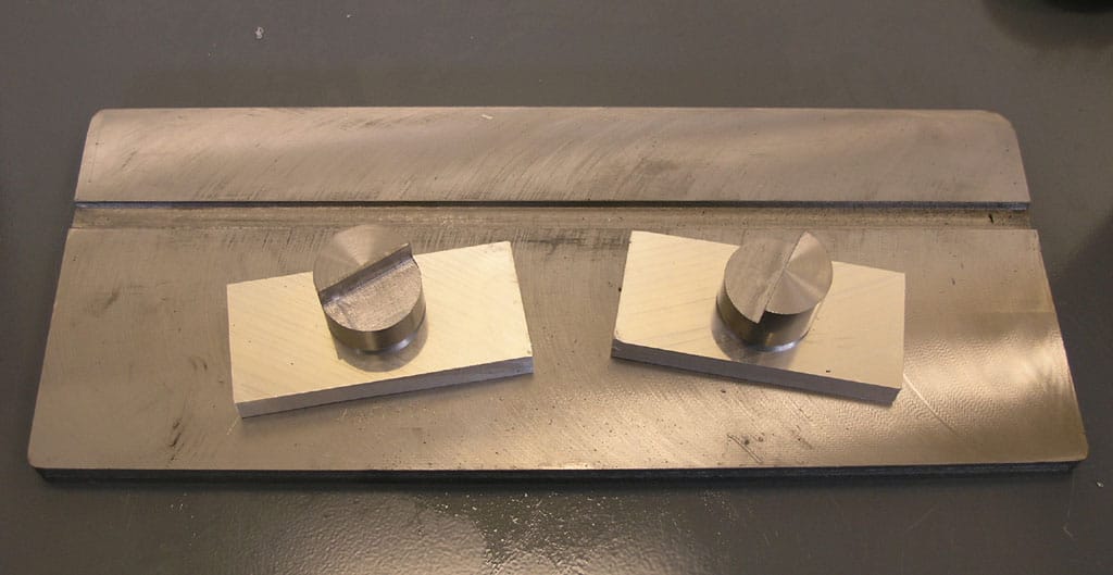 Aluminum bar in vise between two wooden squares with small metal cylinder being pressed by flat metal tool.