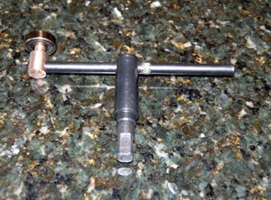 A metal tool with a long handle and round, flat head on one end, featuring a cylindrical body with a threaded end, on a granite countertop.