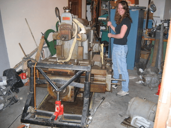 A woman stands in front of a large industrial metal machine surrounded by tools and equipment.