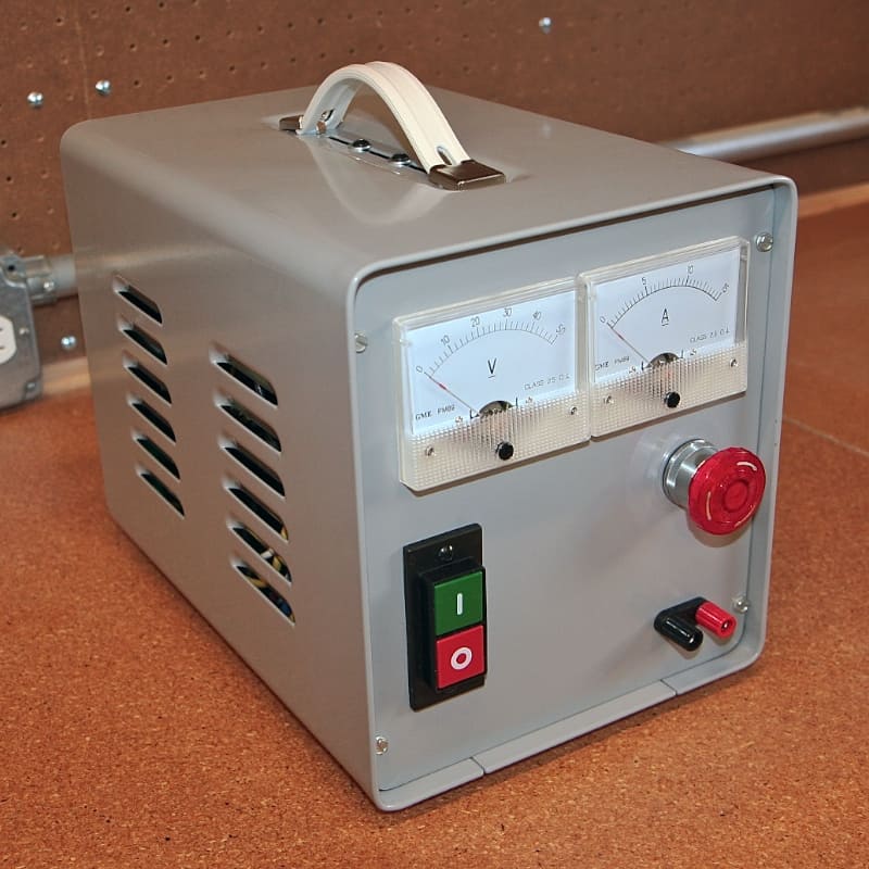 A gray metal enclosure with a handle, featuring two gauges for voltage and current, a red emergency stop button, and a green power switch.