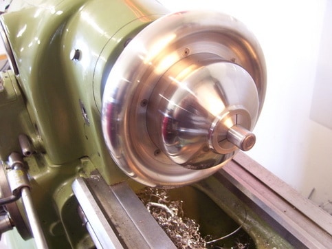 A metal lathe in operation at the home machine shop, showing a spinning chuck and machined part with metal shavings visible.