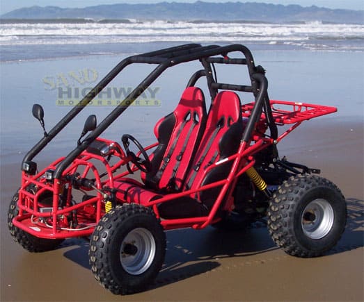 Red mini-sand rail parked on a sandy beach with ocean waves in the background.