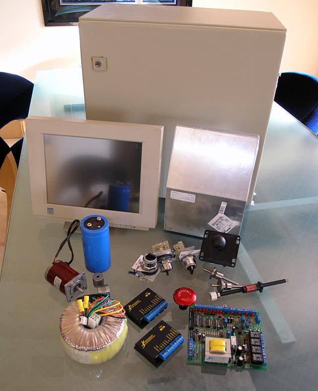 A collection of electronic items including a desktop computer, monitor, camera, and various Lathemaster CNC parts displayed on a glass table.