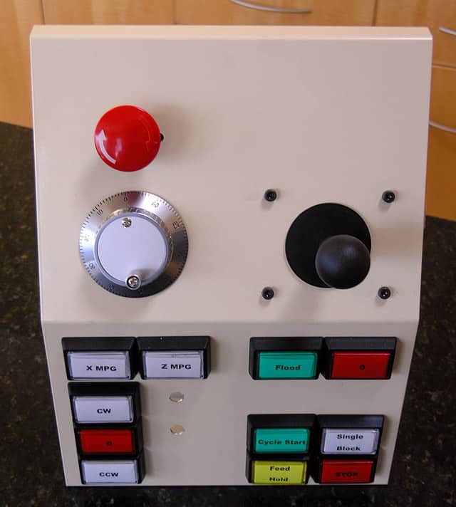 CNC lathe control panel with a red emergency stop button, timer dial, black joystick, and various labeled buttons for machine operations.