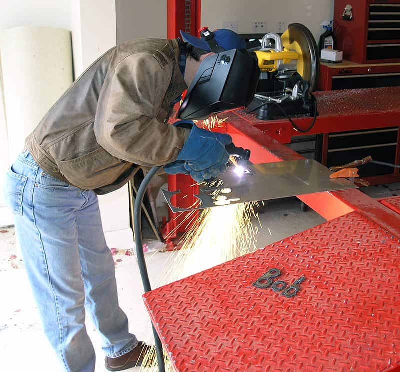 A man welding on a workbench, wearing blue gloves and a black welding helmet with a clear visor.