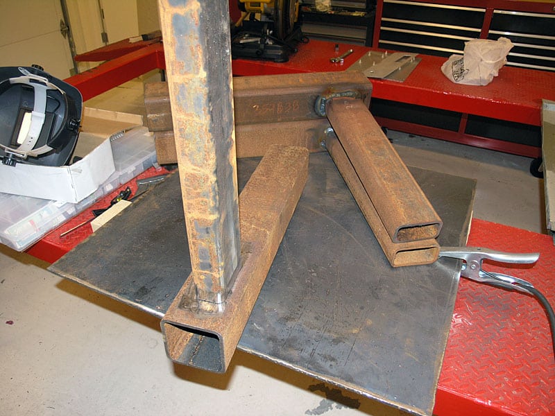 Heavy-duty metal workpiece with three thick steel legs welded to a square base plate, sitting on a red garage floor.