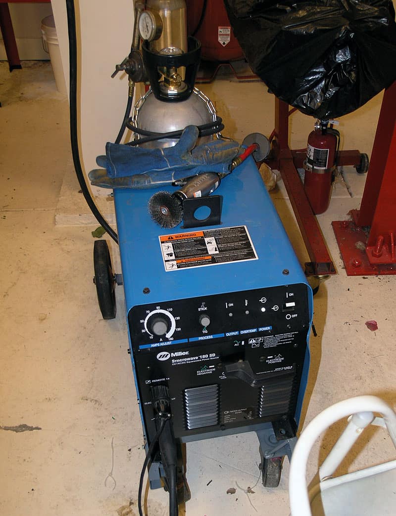 A Miller Syncrowave 180 SD welder in a cluttered workshop environment with various tools and equipment.